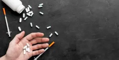 Hand holding pills with pills scattered on table in the background.