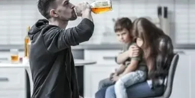 Father drinking alcohol with child and wife in the background.