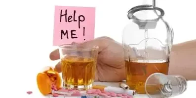 Variety of pills on table with glasses of alcohol and cigarettes. A sign that says help me in the background