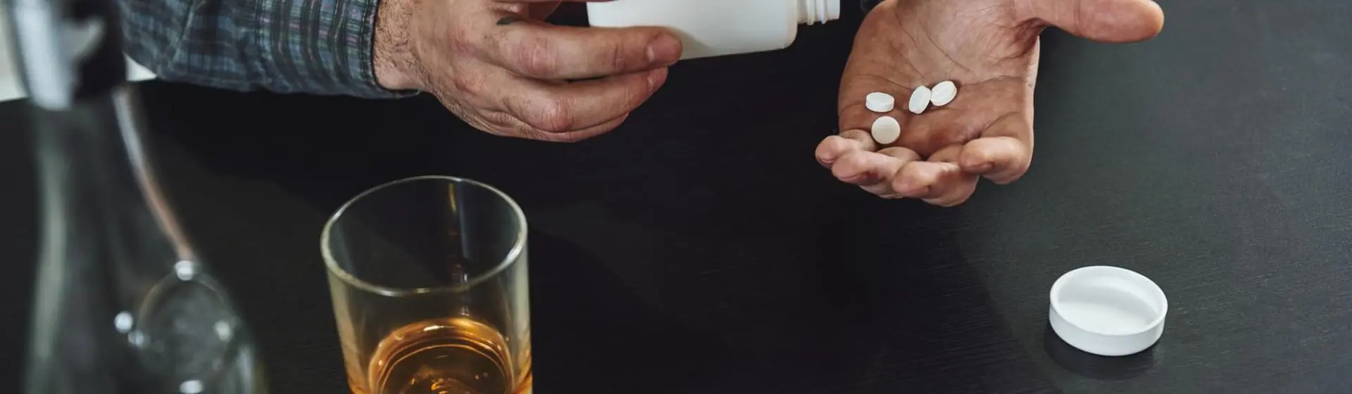 A person pouring pills into one hand with a glass of alcohol on table.