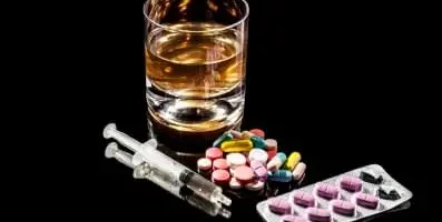 A variety of pills on a table along with a glass of alcohol and syringe.