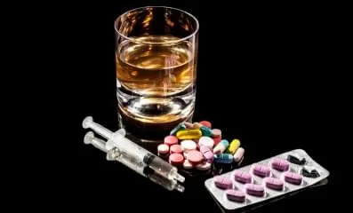 A variety of pills on a table along with a glass of alcohol and syringe.