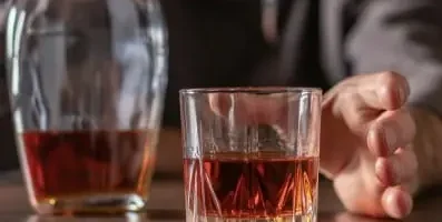 Man reaching for a glass of alcohol on a table. Text: Alcoholism has the potential to escalate, putting the individual at extreme risk.