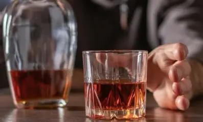 Man reaching for a glass of alcohol on a table. Text: Alcoholism has the potential to escalate, putting the individual at extreme risk.