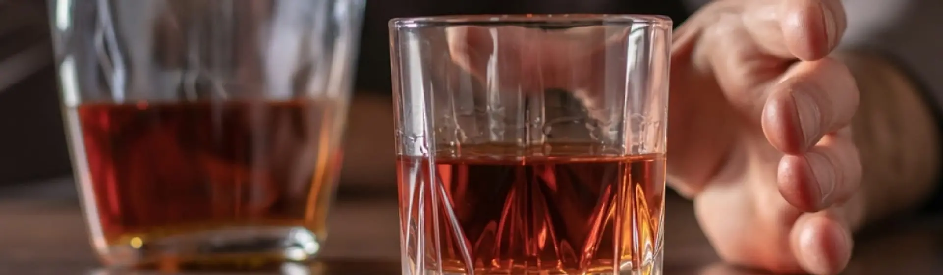 Man reaching for a glass of alcohol on a table. Text: Alcoholism has the potential to escalate, putting the individual at extreme risk.