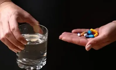 A hand with variety of pills and a glass of water.