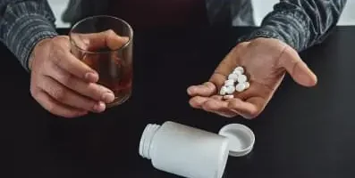 A person holding pills in one hand and glass of alcohol in the other.