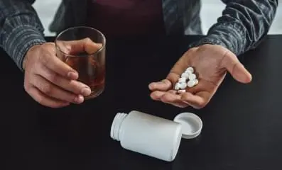 A person holding pills in one hand and glass of alcohol in the other.