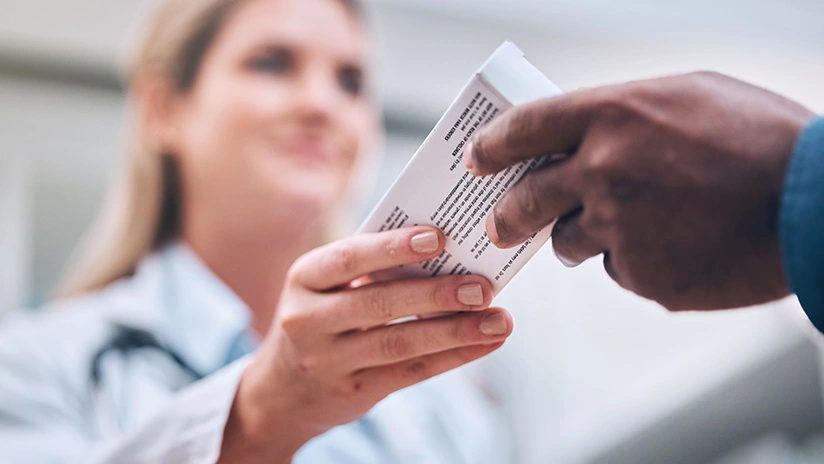 A healthcare professional, likely a doctor, is handing a prescription or informational pamphlet to a patient. The focus is on their hands, with the doctor’s smiling face blurred in the background, suggesting a supportive and professional interaction. The setting appears to be in a clinical or hospital environment.