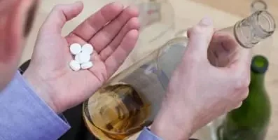 A person holding pills in one hand a bottle of alcohol in the other.