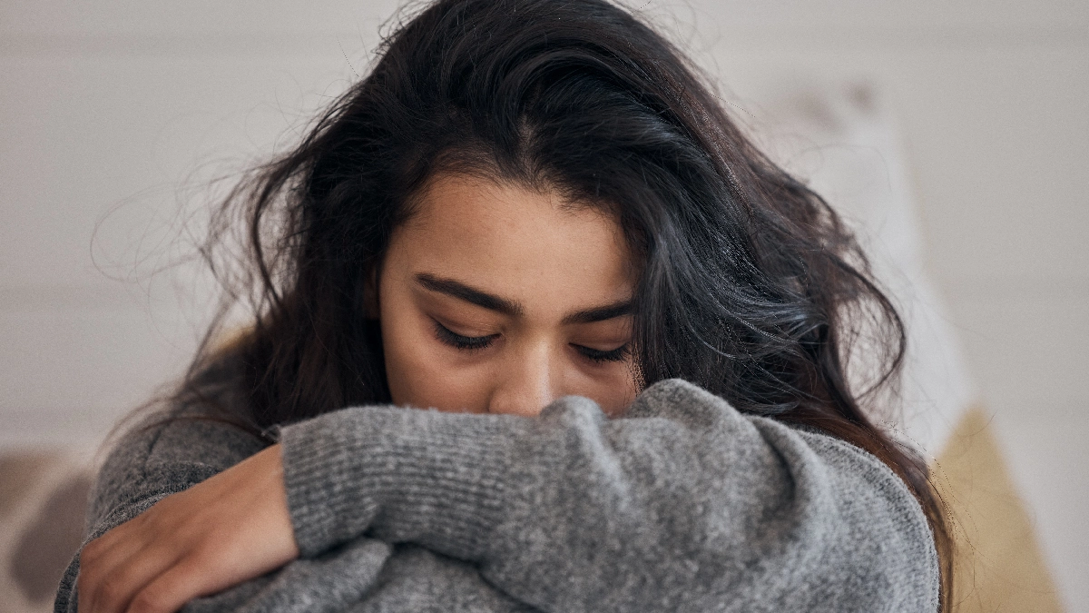 Image of a person with long dark hair, resting their head on their crossed arms and looking down with a sad expression.