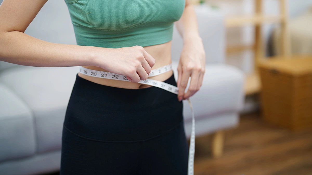 Image of a person in a green crop top measuring their waist with a tape measure.
