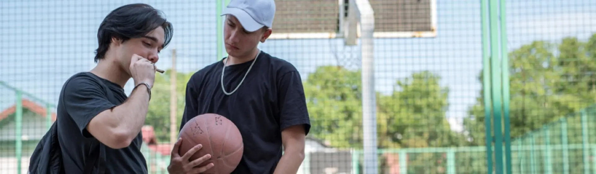 Two teenage boys on an outdoor basketball court, one smokes something.