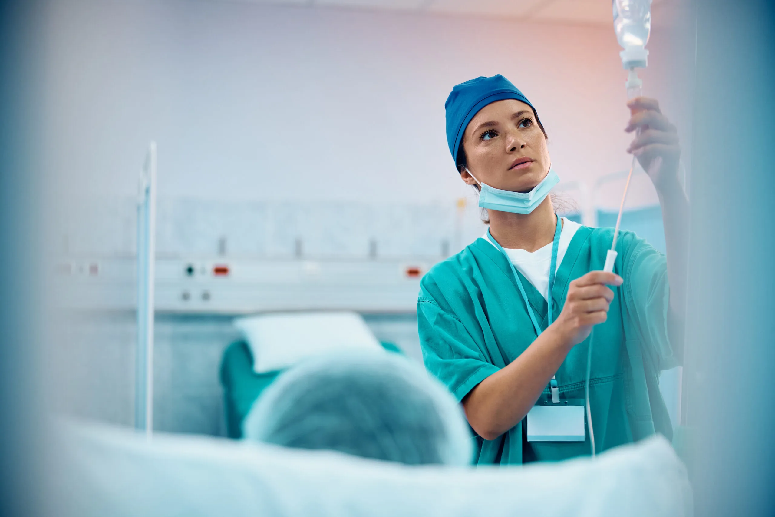 A medical professional in scrubs and a surgical cap is looking at an IV bag while a patient lies in a hospital bed.