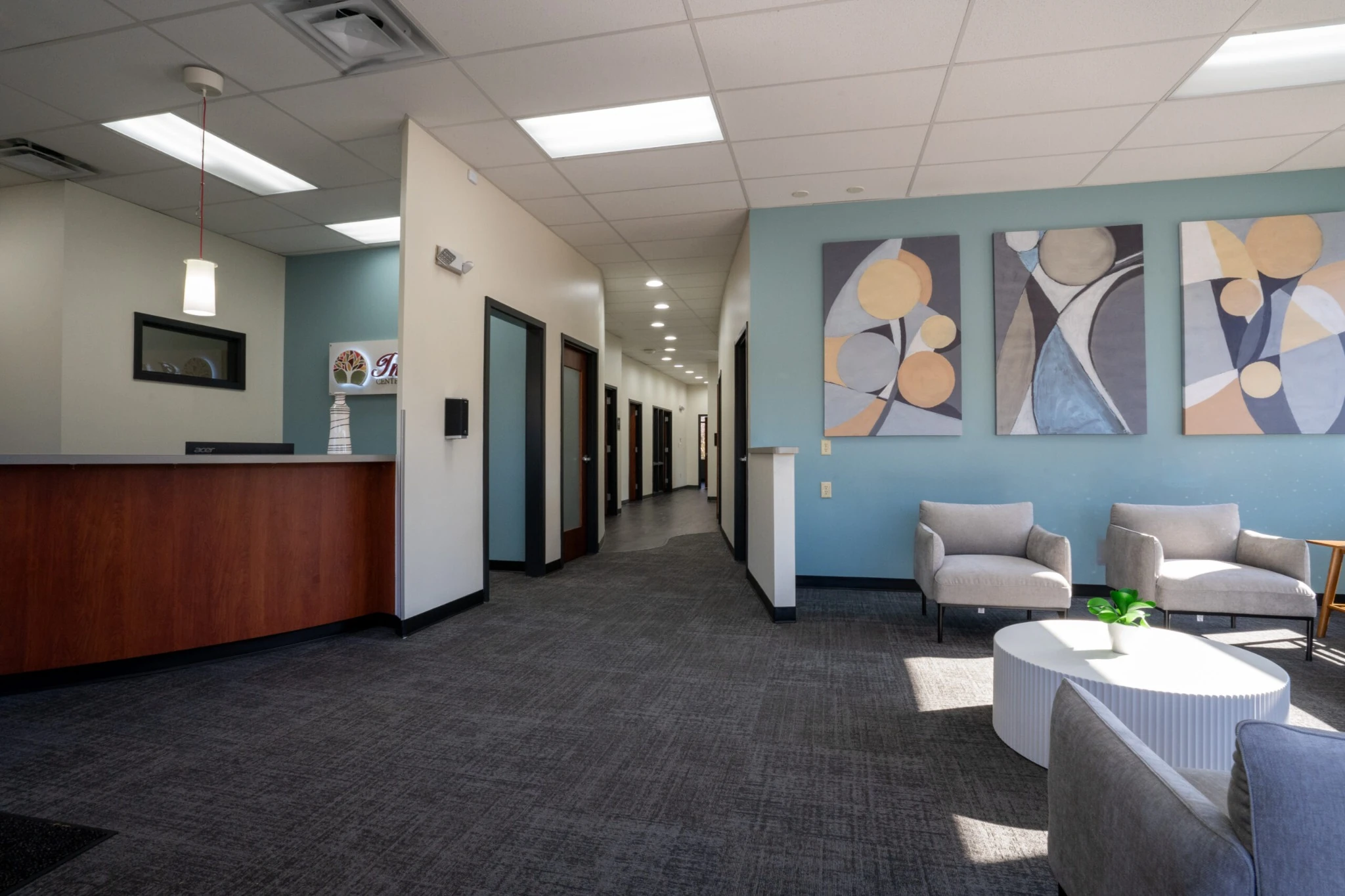 Interior waiting area at Indiana Center for Recovery Carmel with a reception desk on the left, a long hallway of offices, and a seating nook with gray chairs and abstract wall art on a blue accent wall.
