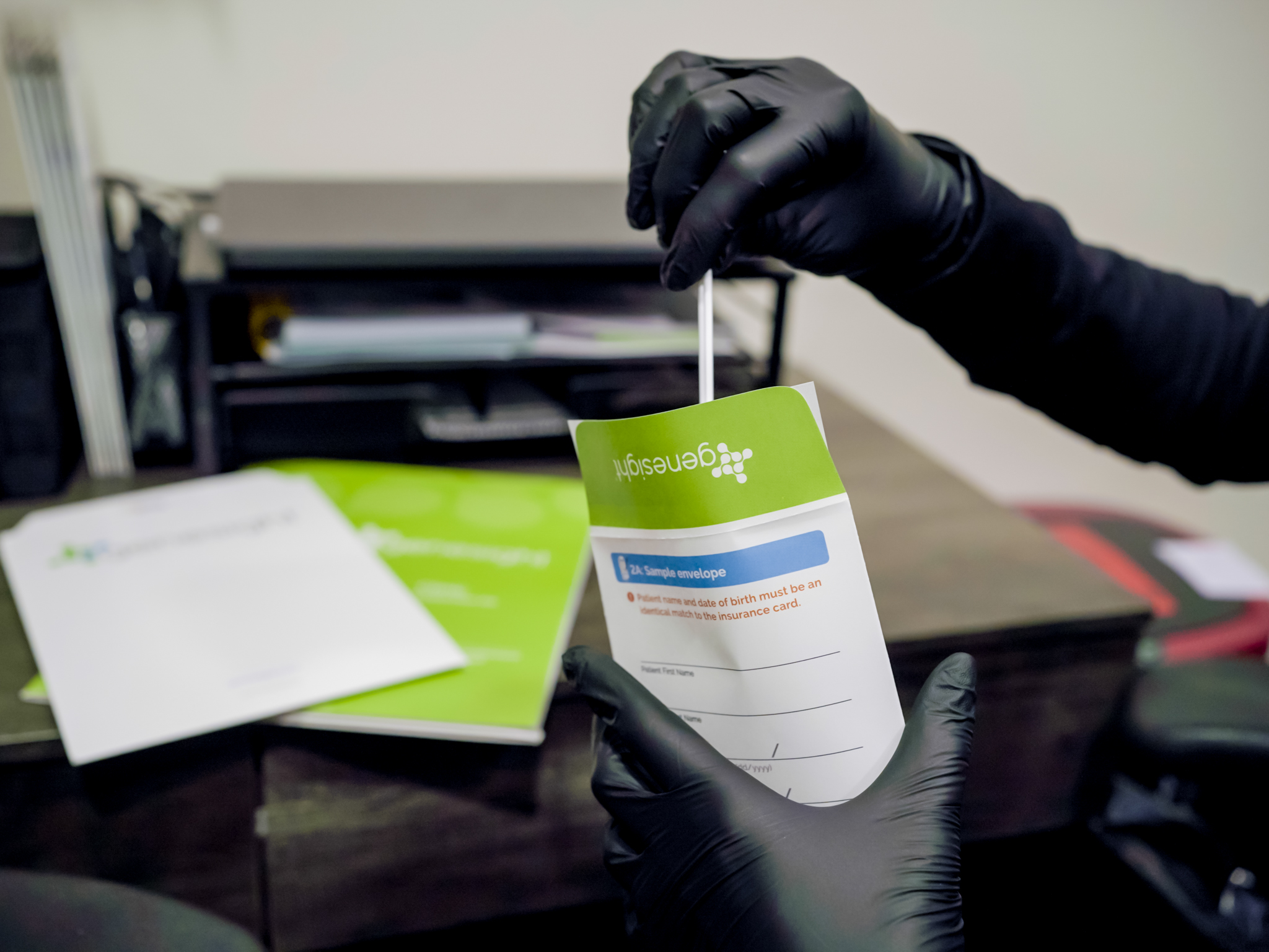 Gloved hands hold a GeneSight testing sample envelope on a desk, with a swab being inserted into the labeled kit and paperwork blurred in the background.