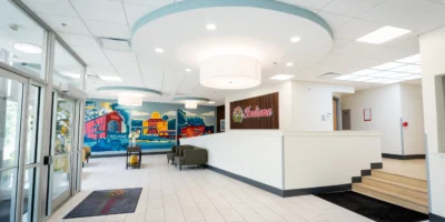 Bright, modern medical lobby with white tile floors, circular ceiling lights, reception desk, seating area, and a colorful Indiana-themed mural.