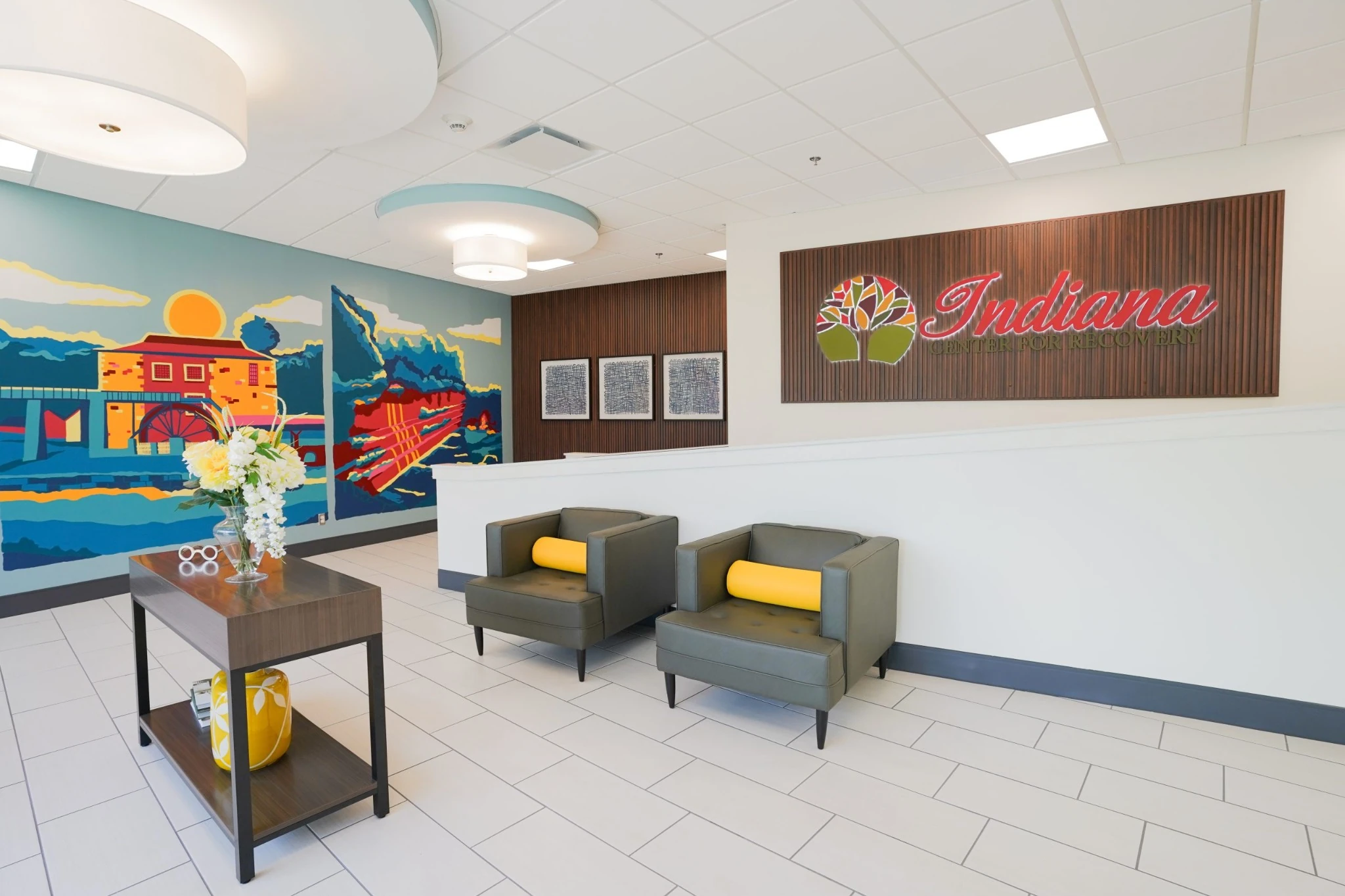 Close-up of Indiana Center for Recovery lobby with reception wall logo, two gray chairs, white tile floors, and a colorful mural.