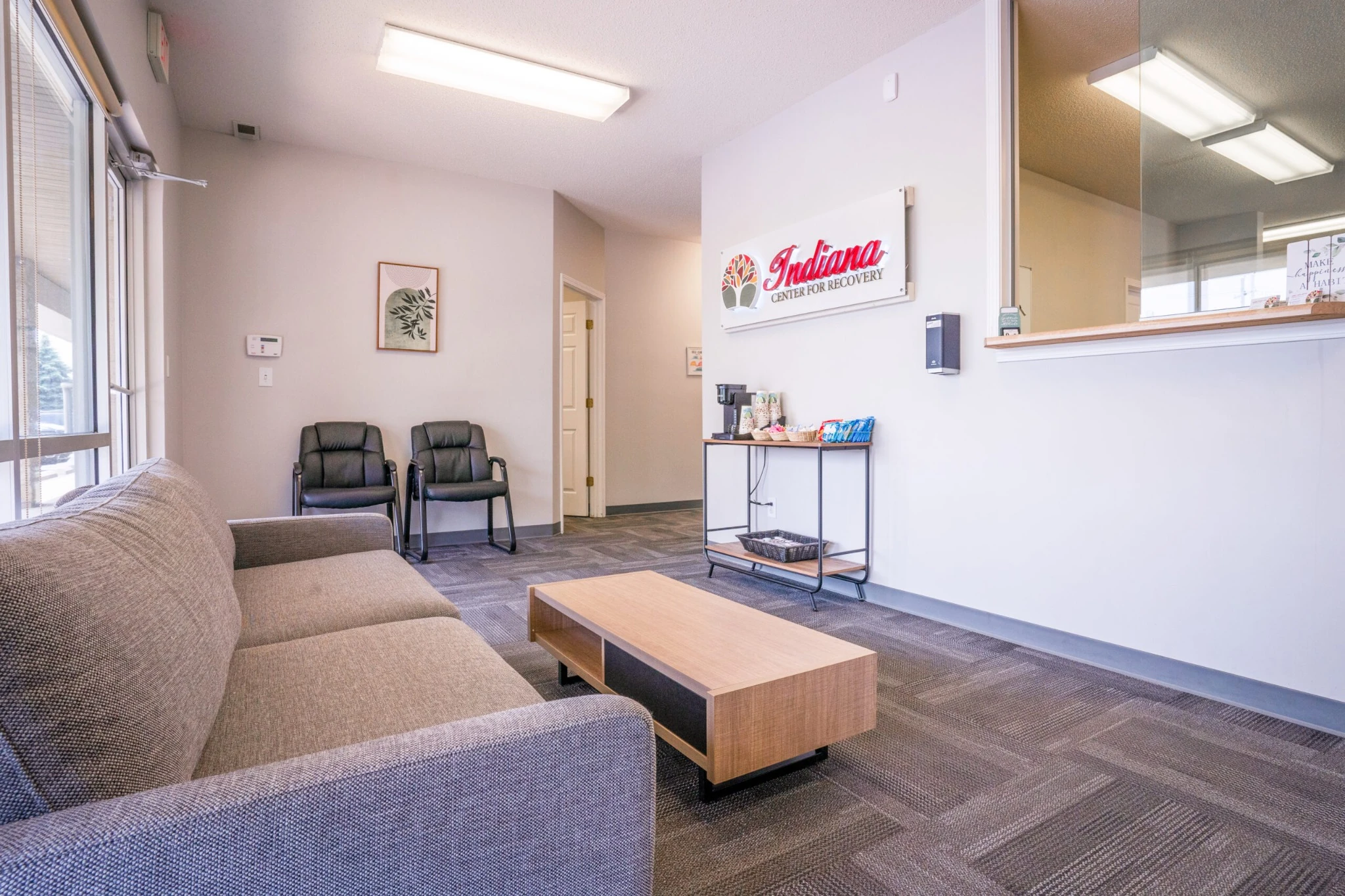 Spacious Indiana Center for Recovery lobby with a gray sofa and light wood coffee table in the foreground, two black chairs against the far wall, a small snack and coffee station under a wall-mounted Indiana Center for Recovery sign, and a reception window on the right.