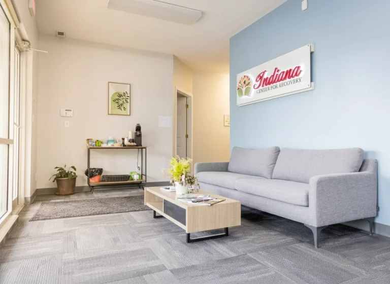 Bright, modern lobby waiting area with a gray sofa on the right, a light wood coffee table with plants and magazines, a small refreshment station on the left, and an ‘Indiana Center for Recovery’ sign mounted on a blue accent wall.