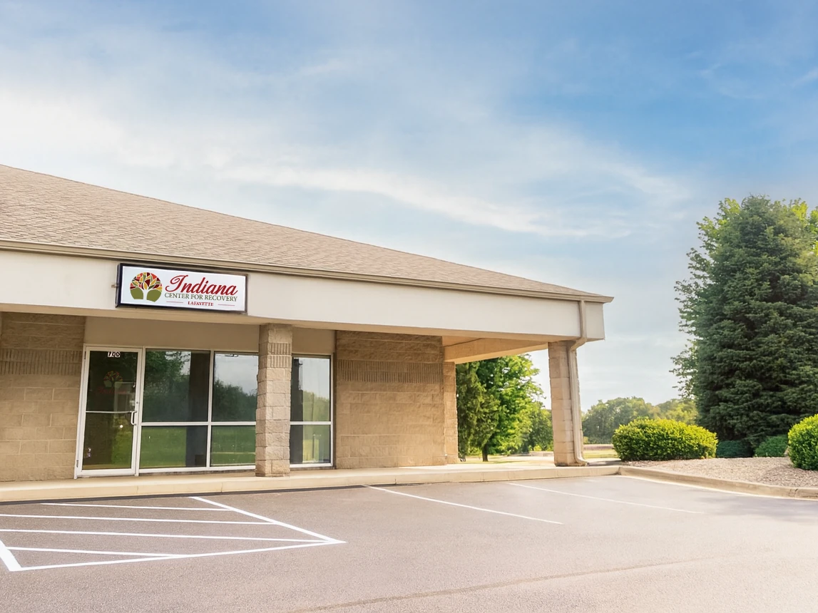 Exterior of Indiana Center for Recovery Lafayette showing a single-story beige building with a covered entrance, ICFR signage above the door, and an empty parking lot on a sunny day.