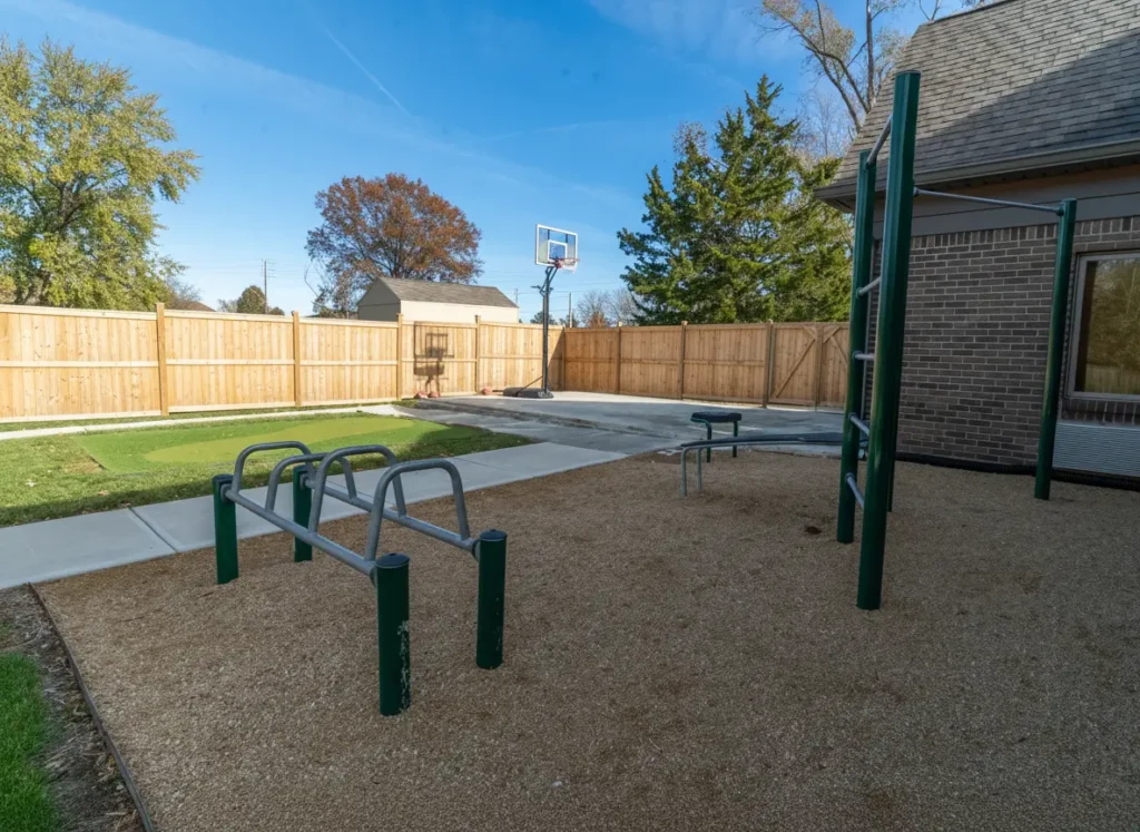 Outdoor recreation area with pull-up bars, parallel bars, and a basketball hoop beside a fenced courtyard under a clear blue sky.