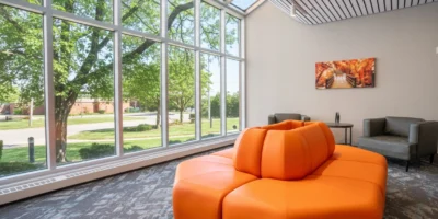 Bright, modern lobby with large floor-to-ceiling windows, an orange modular seating area, and lounge chairs overlooking trees outside.