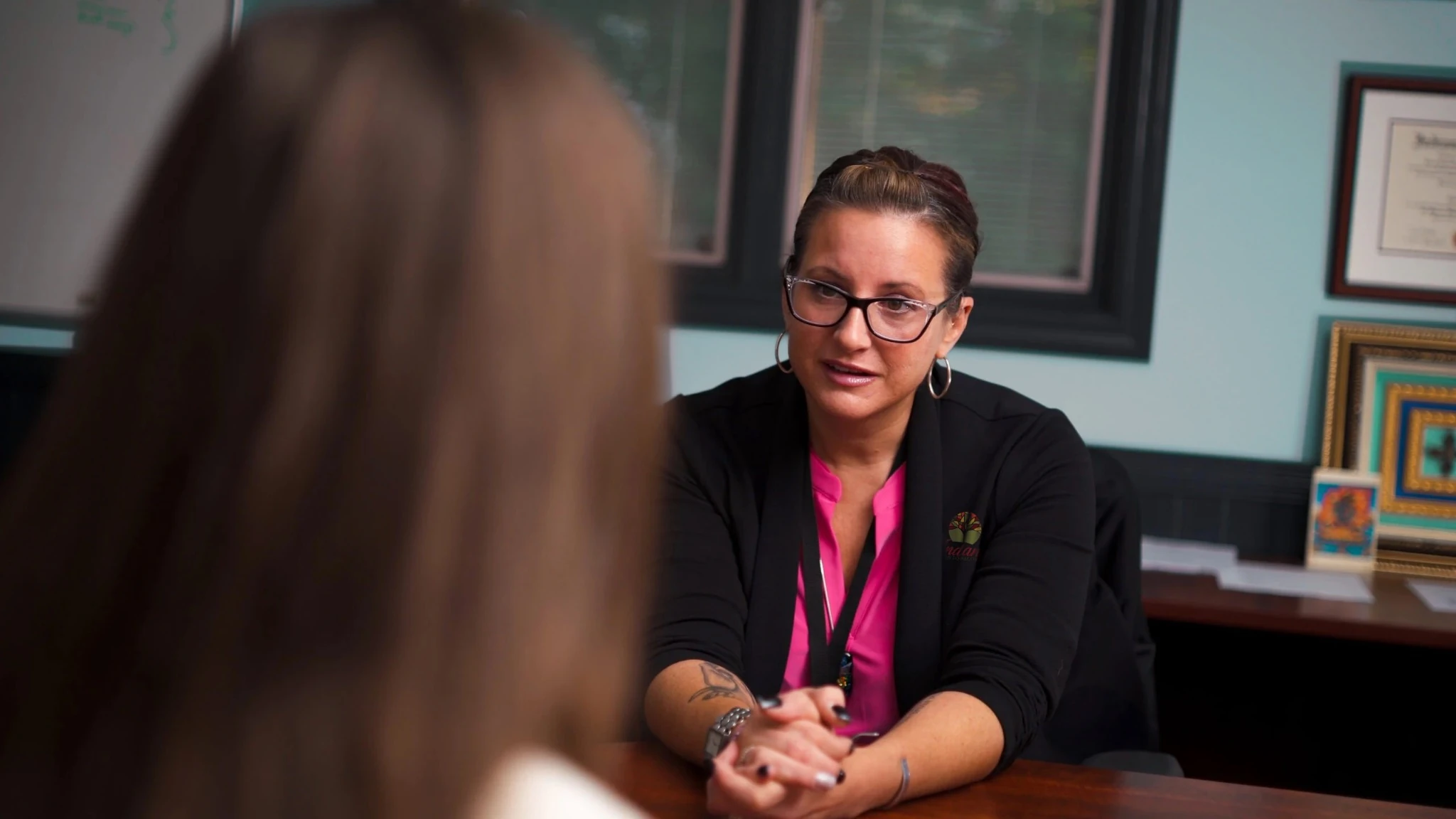 Therapist seated at a desk speaking with a patient in a counseling office, with framed certificates and artwork in the background.