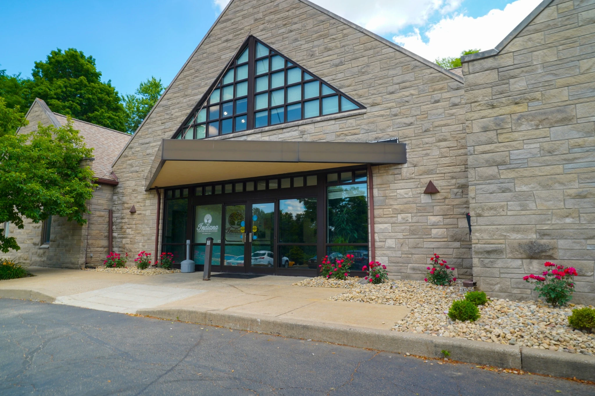 Front entrance of Indiana Center for Recovery Bloomington Walker building with a stone exterior, large triangular window, glass doors, and landscaped flower beds along the walkway.