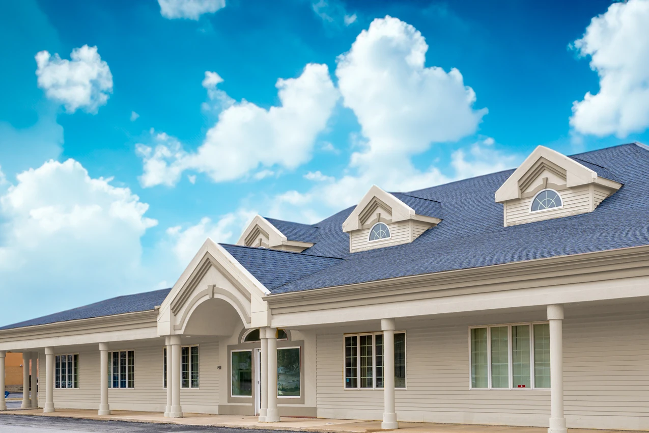 Front exterior of the Indiana Center for Recovery Merrillville facility, a long white building with a covered entrance, columns, and dormer windows under a bright blue sky with clouds.