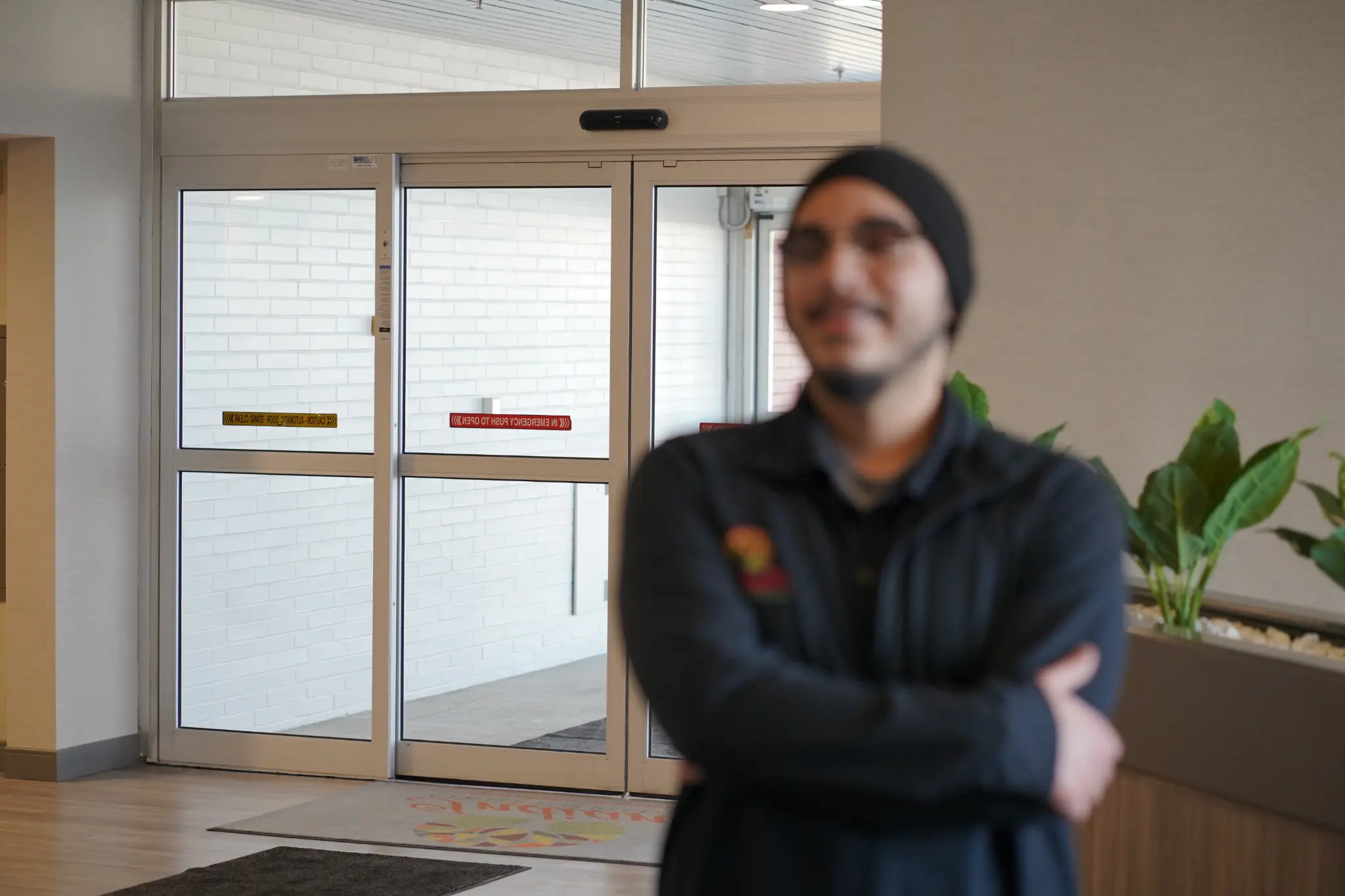 Out-of-focus healthcare staff member standing with arms crossed in a modern medical facility lobby near automatic glass doors.