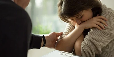 A woman sits with her head down and arms crossed, looking distressed. A person next to her, partially visible, rests a comforting hand on her arm while holding a clipboard, suggesting a supportive or counseling interaction. The setting appears to be in a well-lit room, providing a sense of care and empathy.