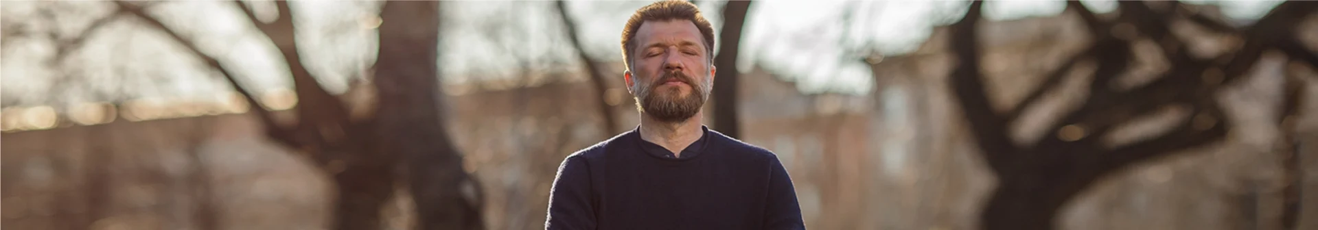 A man with a beard and a dark sweater sits cross-legged on a yoga mat outdoors, meditating with his eyes closed and hands resting on his knees. The background shows a sunny day with trees and blurred scenery.