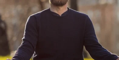 A man with a beard and a dark sweater sits cross-legged on a yoga mat outdoors, meditating with his eyes closed and hands resting on his knees. The background shows a sunny day with trees and blurred scenery.