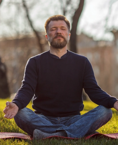 A man with a beard and a dark sweater sits cross-legged on a yoga mat outdoors, meditating with his eyes closed and hands resting on his knees. The background shows a sunny day with trees and blurred scenery.