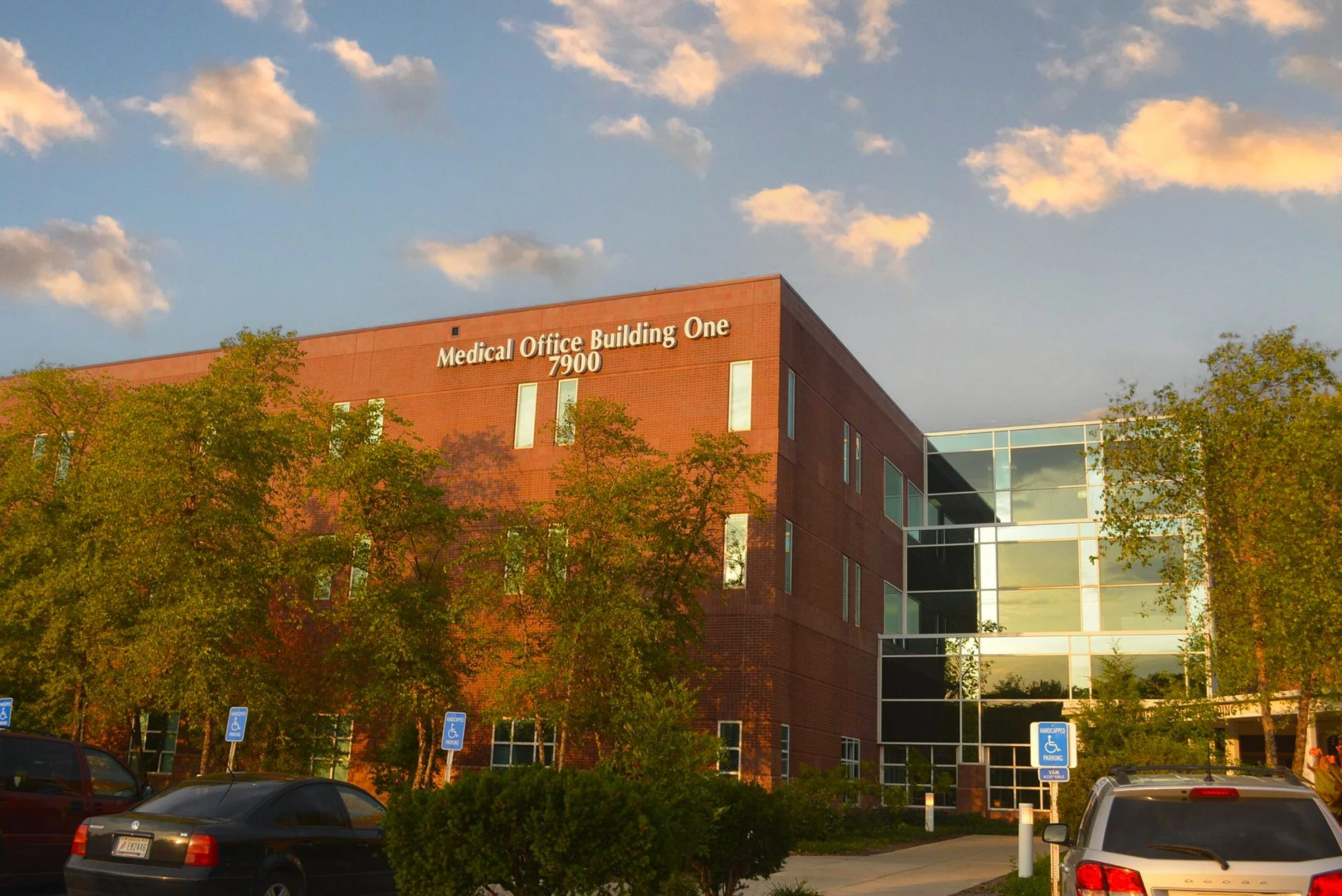 Brick medical office building in Fort Wayne with ‘Medical Office Building One 7900’ signage, large glass entry section, trees in front, and parked cars under a partly cloudy sky at golden hour.