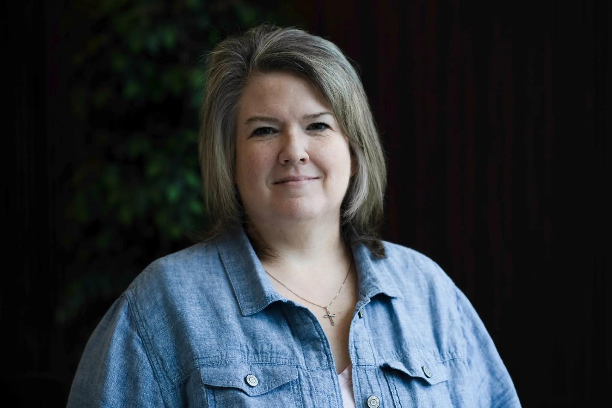 Professional headshot of a woman with shoulder-length gray hair wearing a blue button-up shirt and necklace, photographed against a softly lit, dark background.
