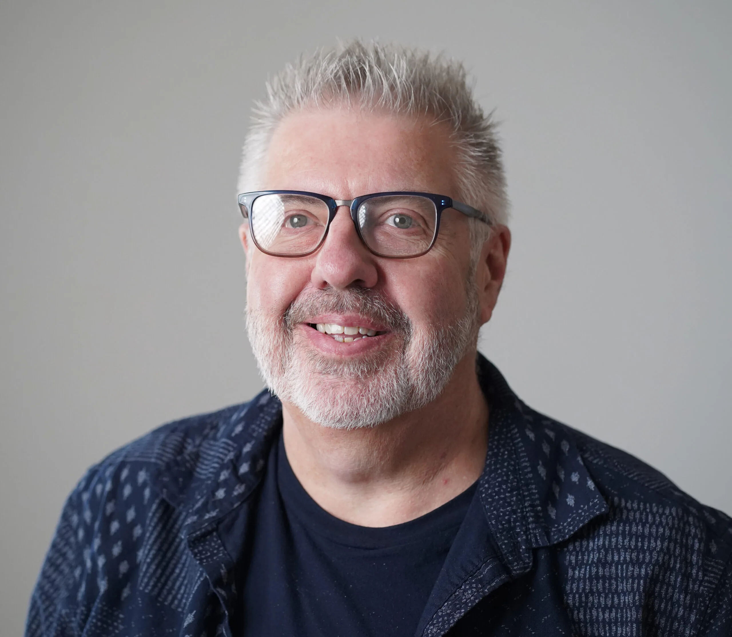 Portrait of a middle-aged man with short gray hair, glasses, and a neatly trimmed gray beard, smiling in a professional headshot against a neutral background, wearing a dark patterned jacket and black shirt.