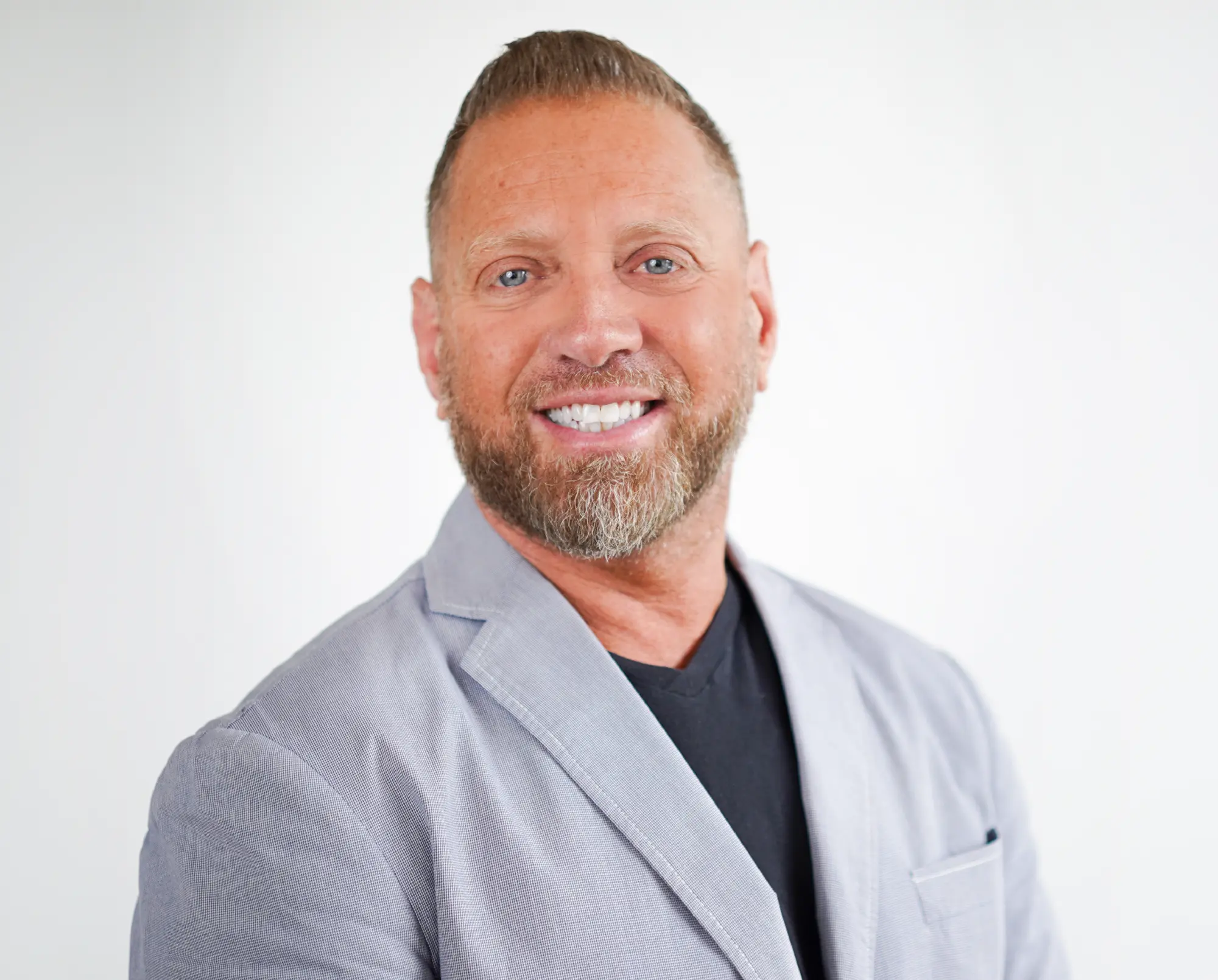 Smiling man with short hair and a trimmed beard, wearing a light gray blazer over a dark shirt against a white background.