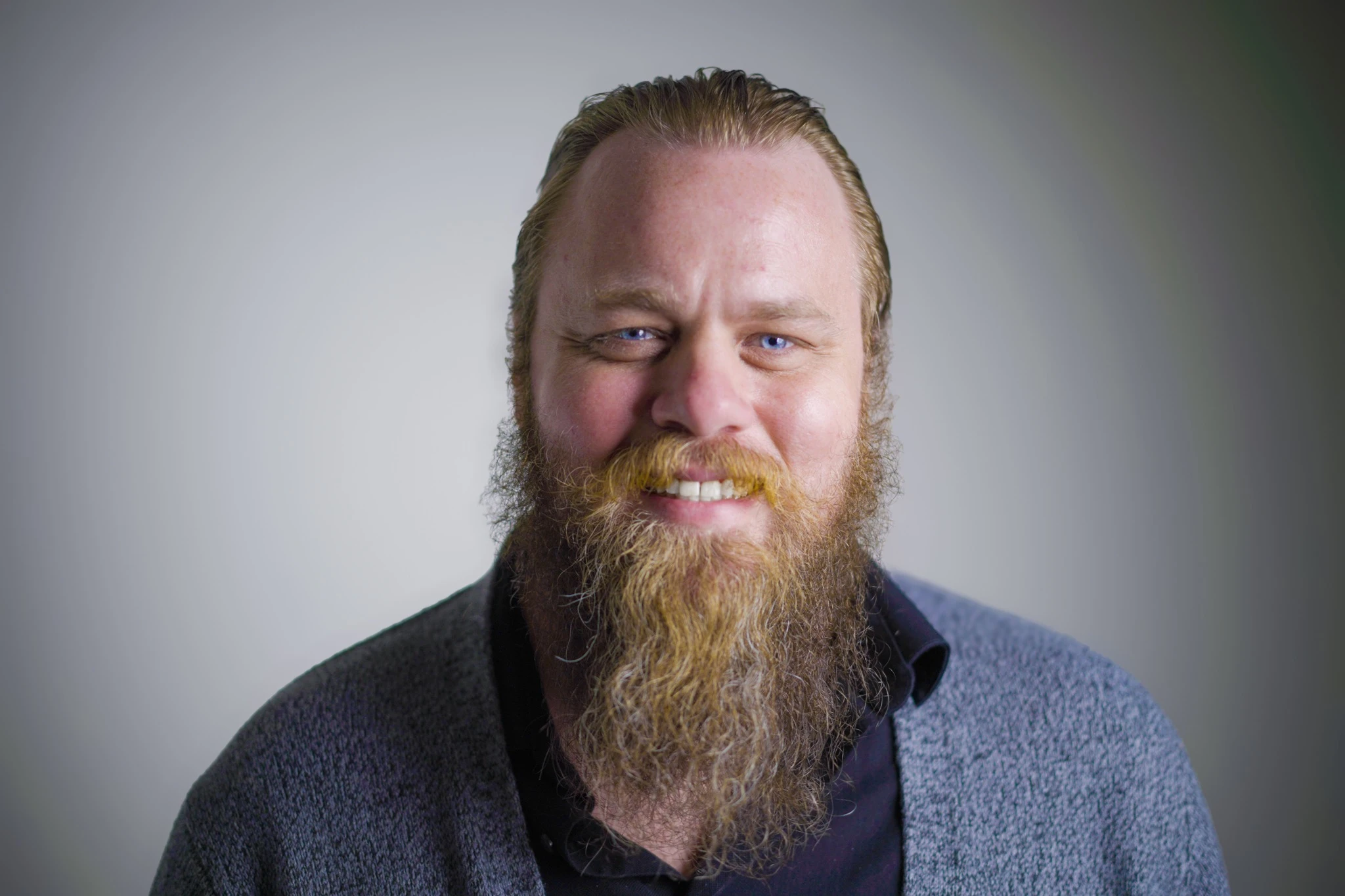 Portrait of a smiling man with slicked-back hair and a long reddish-brown beard, wearing a dark shirt and gray sweater against a neutral background.