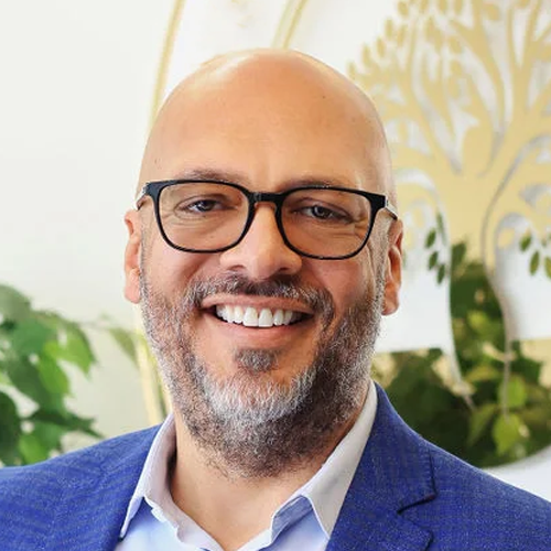 Portrait of Carl Ratcliff smiling in a blue blazer and glasses, professional healthcare leadership headshot