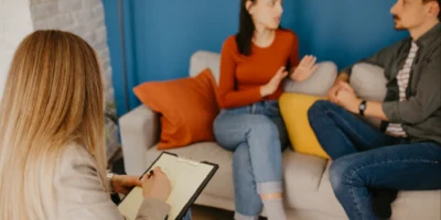 Therapist taking notes while a couple sits on a couch discussing concerns during a counseling session.