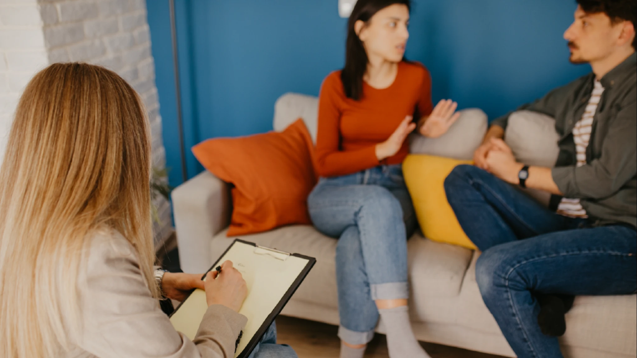 Therapist taking notes while a couple sits on a couch discussing concerns during a counseling session.