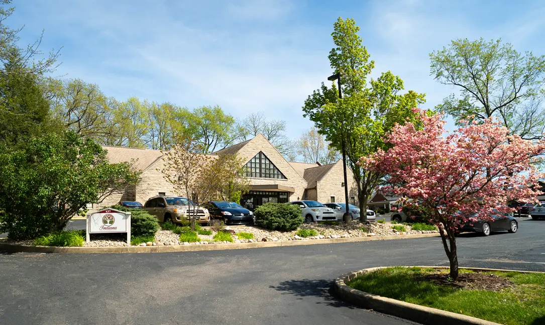 Exterior view of an Indiana Center for Recovery building surrounded by trees and landscaping, with parked cars in front and a blooming pink tree along the driveway on a sunny day.