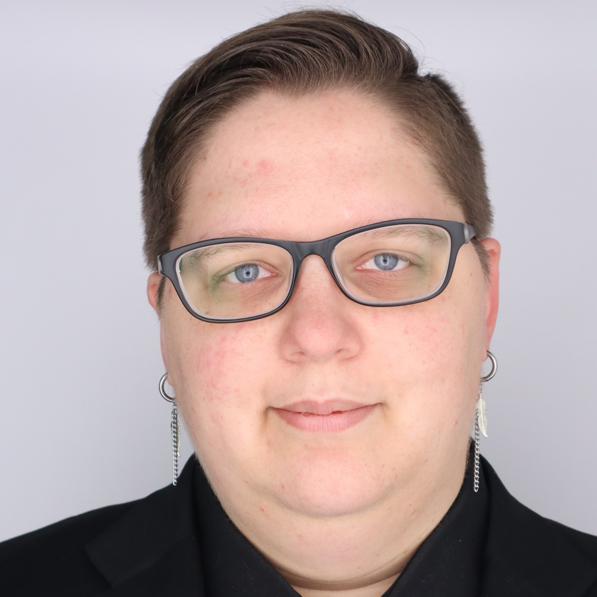 Professional headshot of a staff member wearing black-framed glasses, short hair, and a black blazer against a light gray background