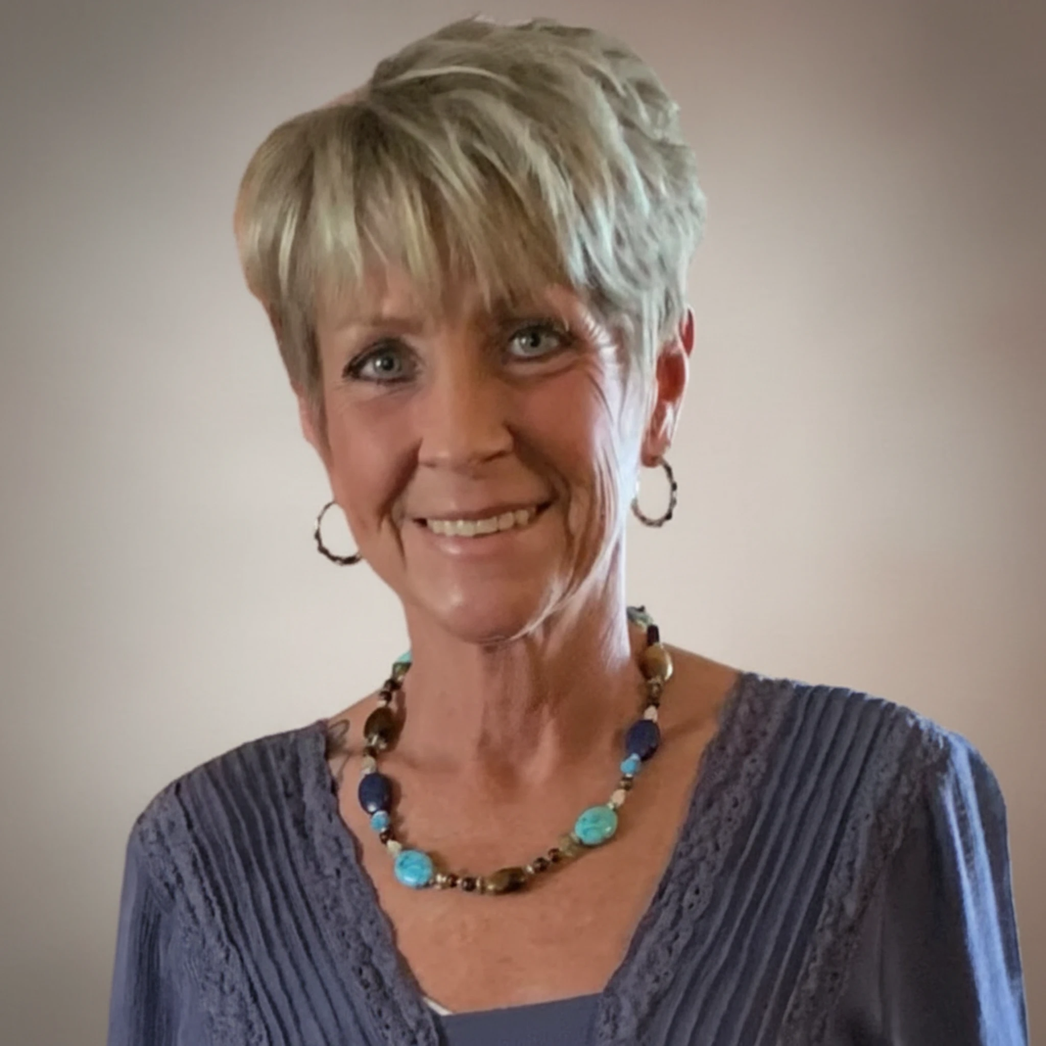 Portrait of a smiling woman with short blonde hair wearing hoop earrings, a blue blouse, and a multicolored beaded necklace against a neutral background.