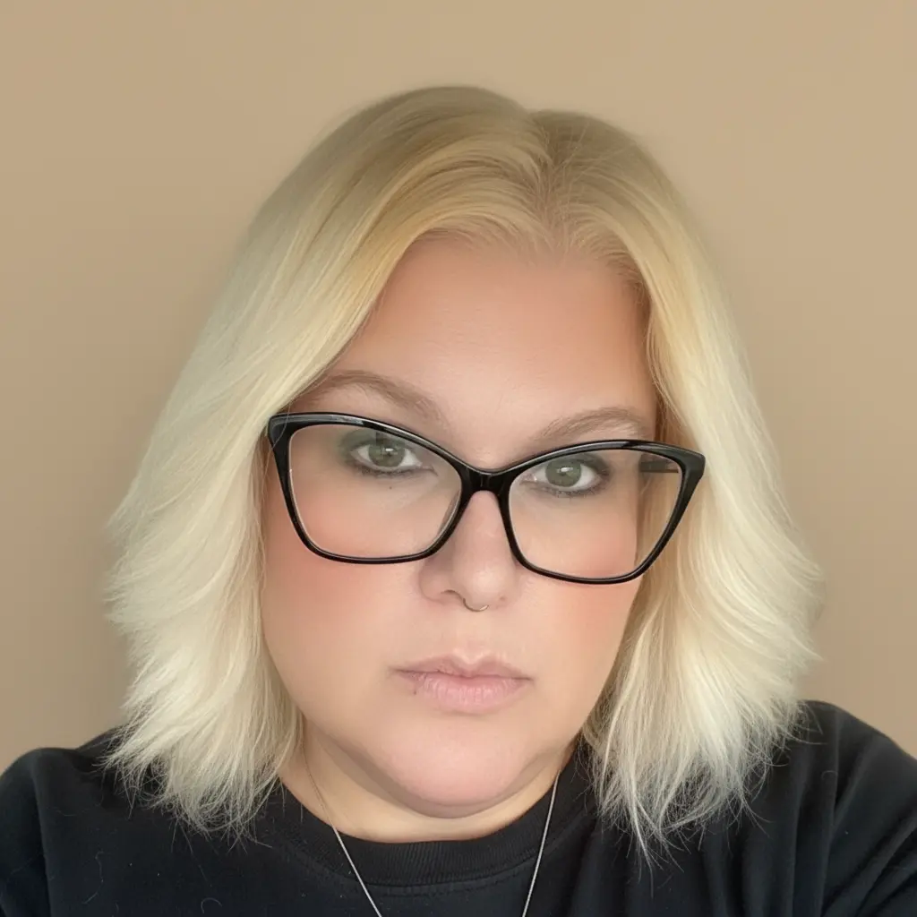 ortrait of a woman with shoulder-length blonde hair wearing black-framed glasses and a black top, looking directly at the camera against a neutral background.