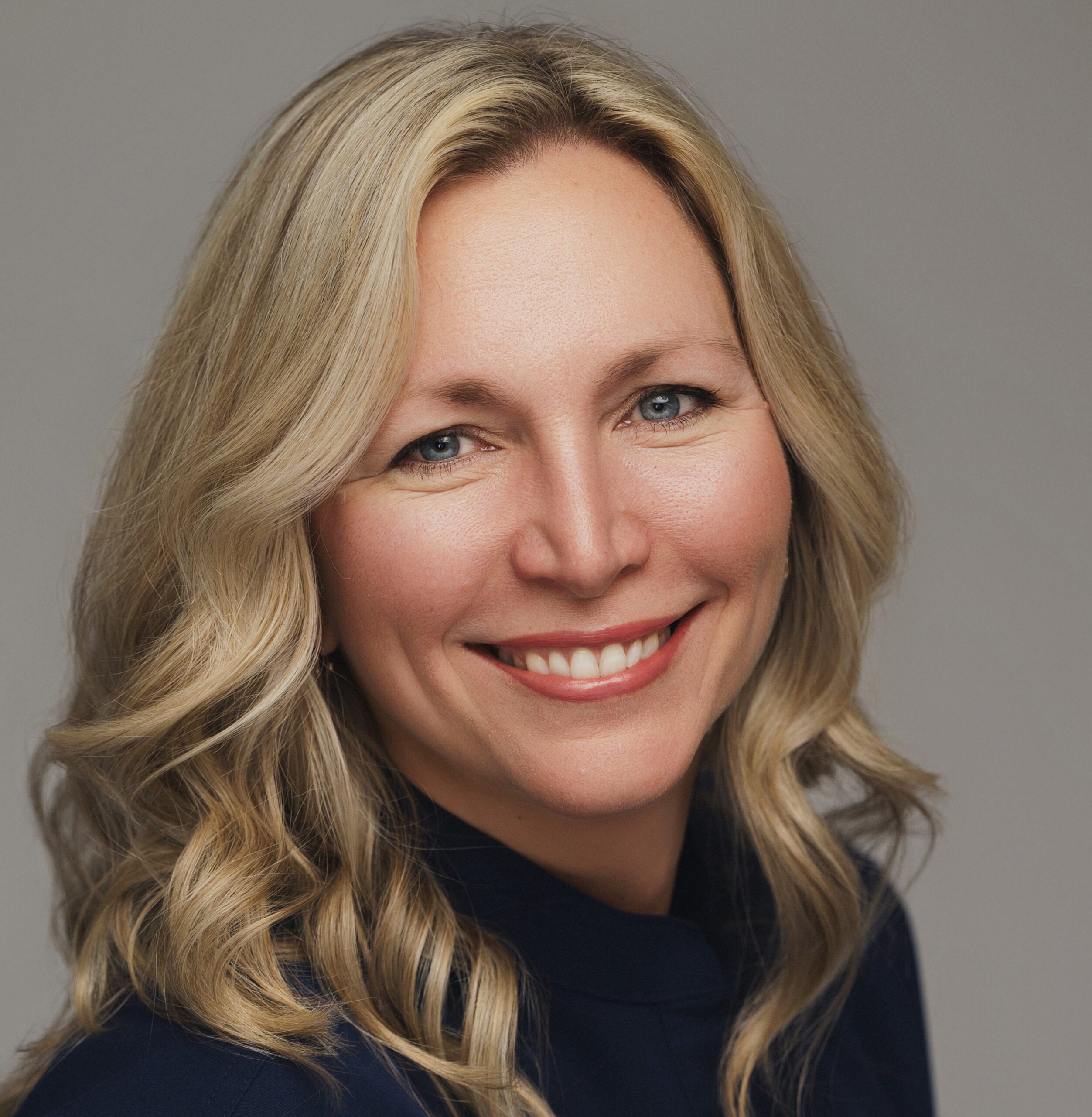 Professional headshot of a smiling woman with long blonde hair wearing a dark top against a neutral gray background.
