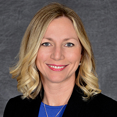 Professional headshot of a smiling woman with shoulder-length blonde hair, wearing a black blazer over a blue top, against a gray textured background.