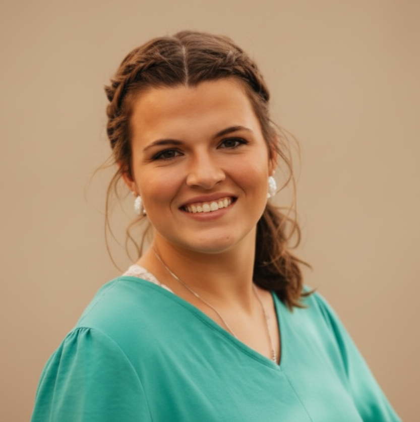 Smiling woman with braided hair pulled back in a ponytail, wearing a teal V-neck blouse and earrings, posed outdoors against a softly blurred neutral background.